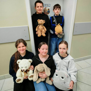 Trzy dziewczyny siedzą na podłodze, dwóch chłopców stoi za nimi w drzwiach do klasy, wszyscy trzymają maskotki.